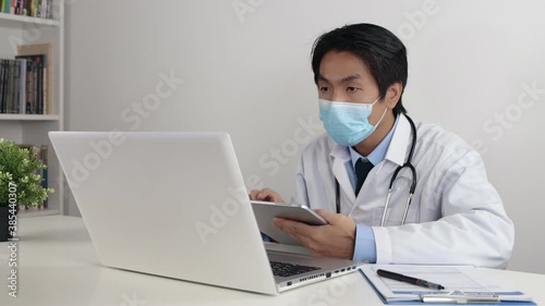 Young Asian Doctor Man in Lab Coat or Gown with Stethoscope Wear Face Mask Check Physical Examination Report from Tablet and Using Laptop in Office