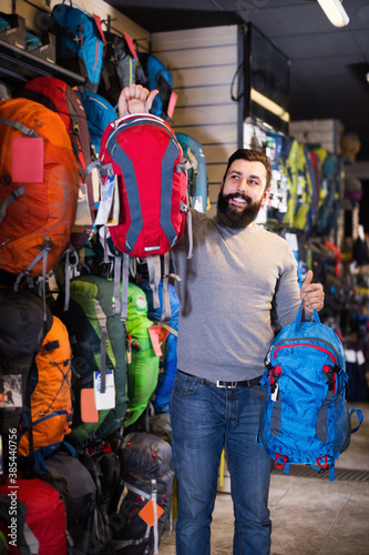Smiling man customer examining backpacks in sports equipment store.