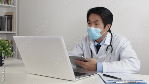 Young Asian Doctor Man in Lab Coat or Gown with Stethoscope Wear Face Mask Exam Patient by Tablet and Laptop via Video Chat or Video Conference in Office