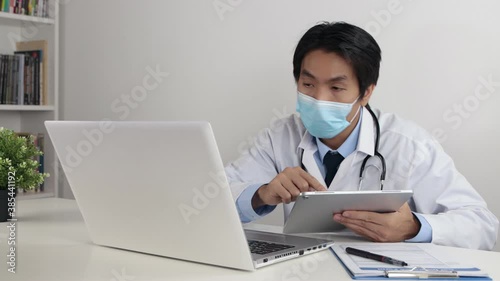 Young Asian Doctor Man in Lab Coat or Gown with Stethoscope Wear Face Mask Online Meeting or Video Conference by Tablet and Laptop in Office