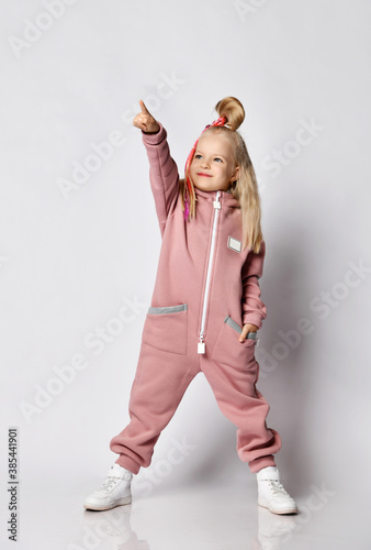 Cool blonde 6-7 y.o. kid girl with girlish hairstyle in pink jumpsuit stands pointing with finger at upper corner