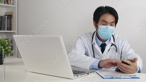 Young Asian Doctor Man in Lab Coat or Gown with Stethoscope Wear Face Mask Using Smartphone and Writing Physical Examination Report in Office