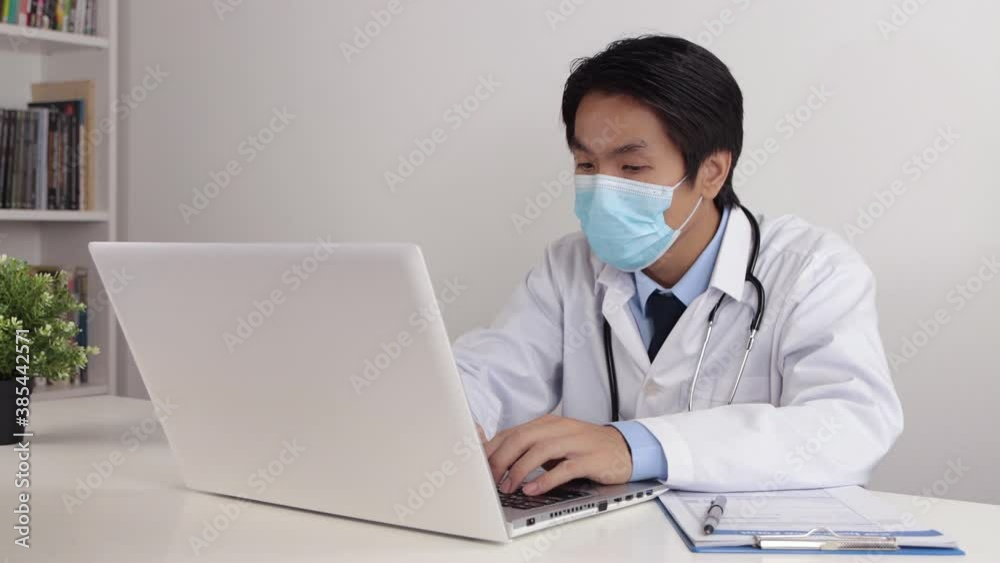 Young Asian Doctor Man in Lab Coat or Gown with Stethoscope Wear Face Mask Using Laptop Computer on Doctor Table in Office Young Asian Doctor Man in Lab Coat or Gown with Stethoscope Wear Face Mask Using Laptop Computer on Doctor Table in Office