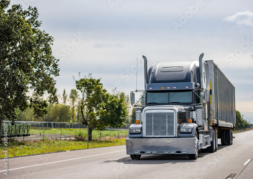 Black big rig classic semi truck with chrome accessories transporting cargo in long industrial container running on the straight highway