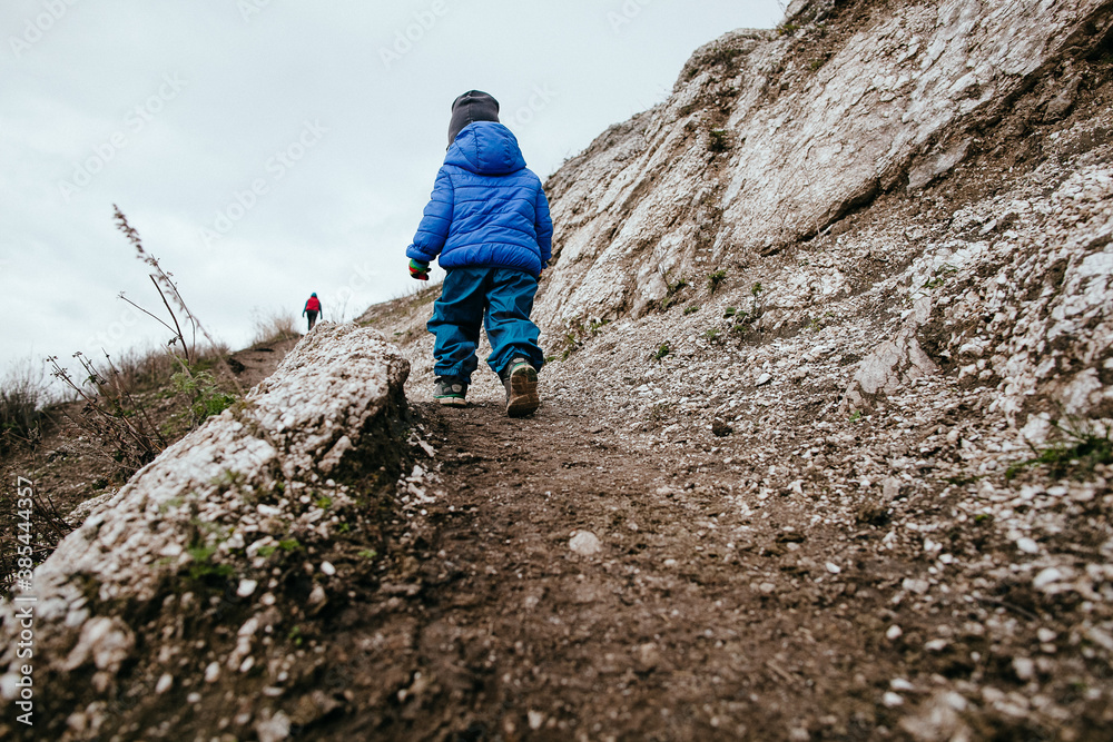 A child in a blue jacket walks along the path. A little boy climbs the mountain on his own. Children's outdoor trekking in nature A child in a blue jacket walks along the path. A little boy climbs the mountain on his own. Children's outdoor trekking in nature