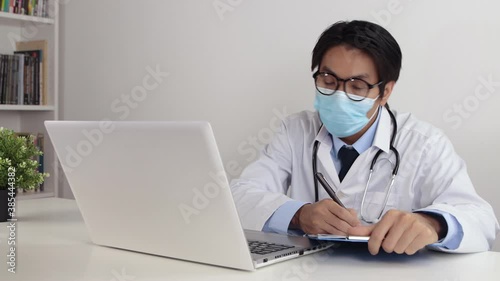 Young Asian Doctor Man in Lab Coat or Gown with Stethoscope Wear Glasses and Face Mask Exam Patient by Video Call or Video Conference in Office