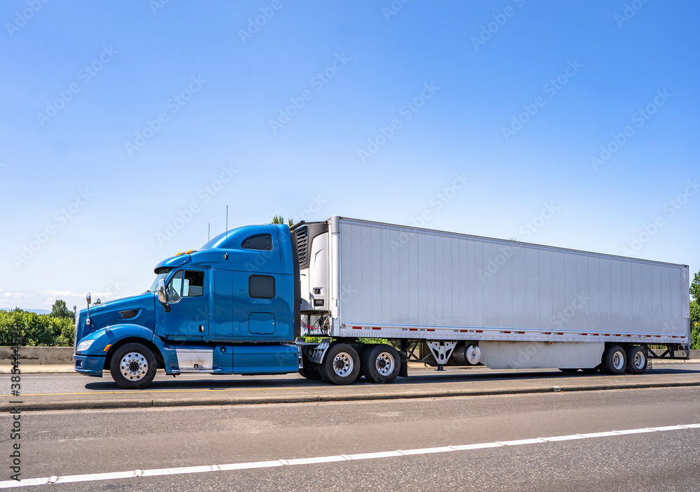 Side of blue big rig semi truck tractor transporting frozen cargo in refrigerator semi trailer driving on the wide multiline highway road