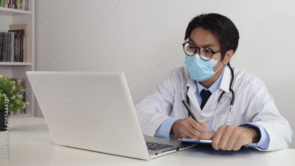 Young Asian Doctor Man in Lab Coat or Gown with Stethoscope Wear Glasses and Face Mask Suggest and Exam Patient by Video Call or Video Conference in Office Young Asian Doctor Man in Lab Coat or Gown with Stethoscope Wear Glasses and Face Mask Suggest and Exam Patient by Video Call or Video Conference in Office