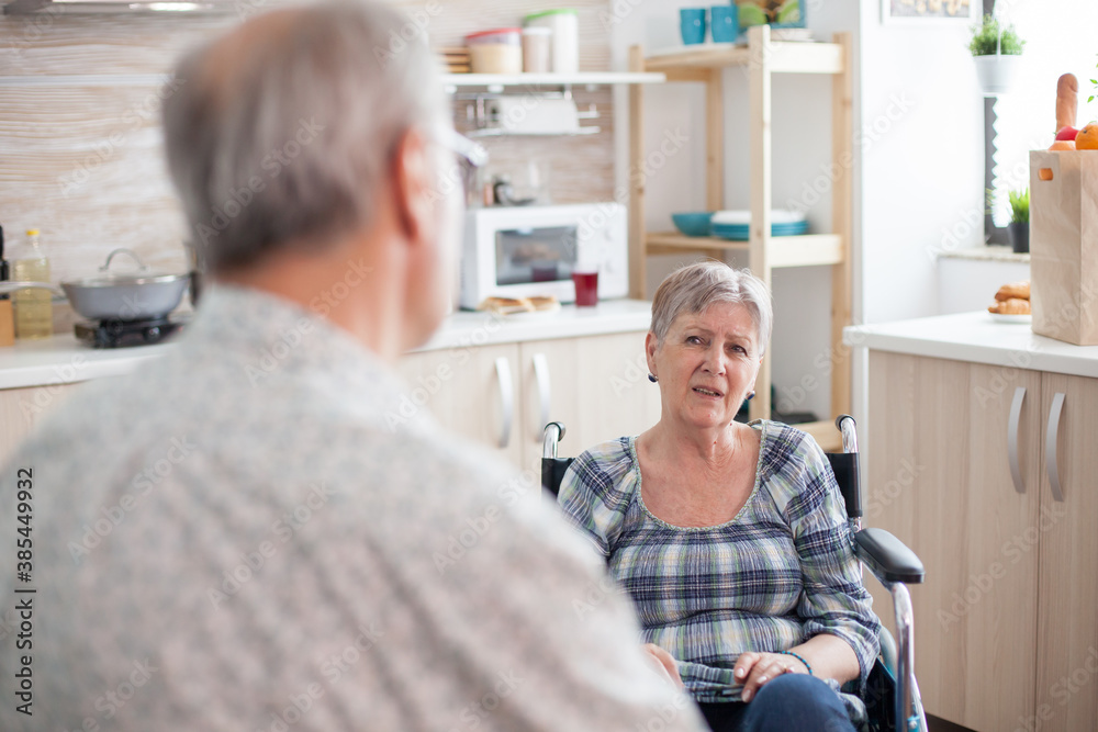 Obraz premium Gray Haired man chatting with paralysed wife. Retired invalid woman in wheelchair having a conversation with old elderly husband in kitchen. Old man talking with wife. Living with disabled person with