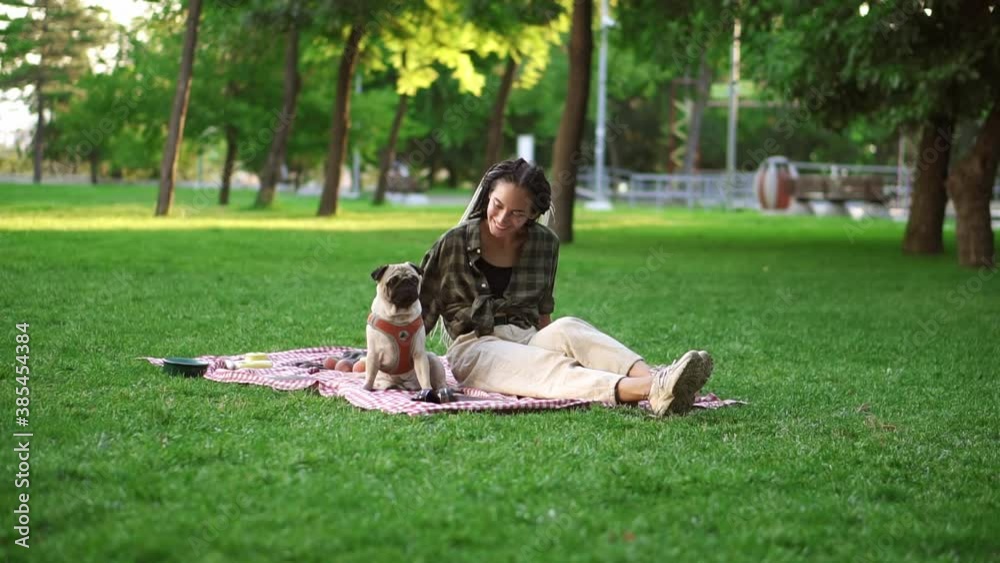 Smiling girl sitting on plaid on lawn in a park., the dog sitting in next to her, she is wondering, looking at him Smiling girl sitting on plaid on lawn in a park., the dog sitting in next to her, she is wondering, looking at him