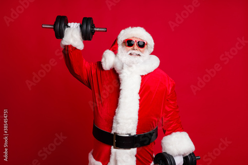 Active life for pensioners. Photo of retired old man white beard lift heavy dumbbells smiling strong grandpa concept wear santa costume sunglass headwear isolated red color background