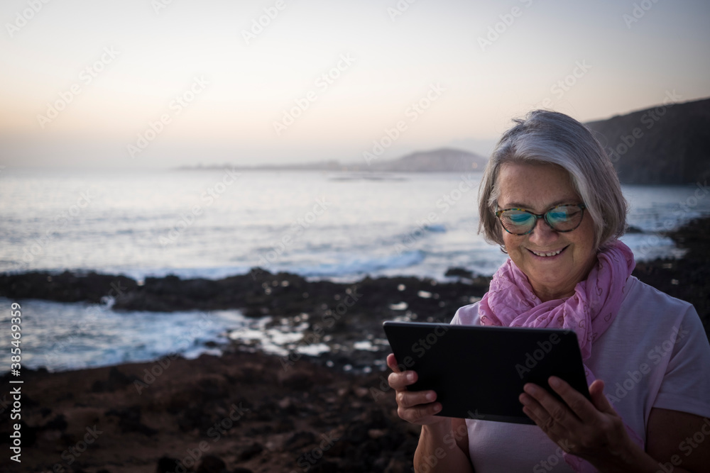 A smiling senior woman sitting near the beach with dusk light looking at the tablet in video call with friends. Happy retirement concept. Sea in the background