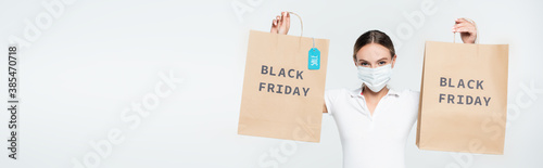 panoramic shot of customer in medical mask holding shopping bags with sale tag and black friday lettering isolated on white