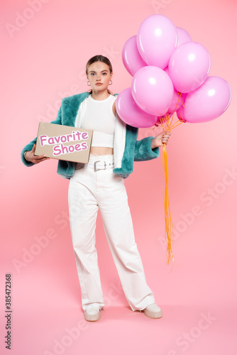young woman holding carton box with favorite shoes lettering and balloons while standing on pink