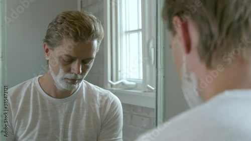 Reflection of a blonde haired man shaves off a scraggily beard with a safety razor after quarantine. photo