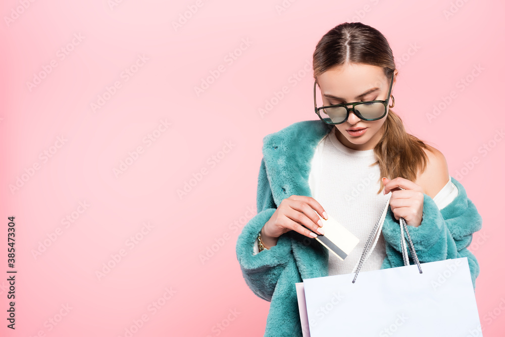 young trendy woman in sunglasses holding credit card near shopping bag isolated on pink, black friday concept young trendy woman in sunglasses holding credit card near shopping bag isolated on pink, black friday concept