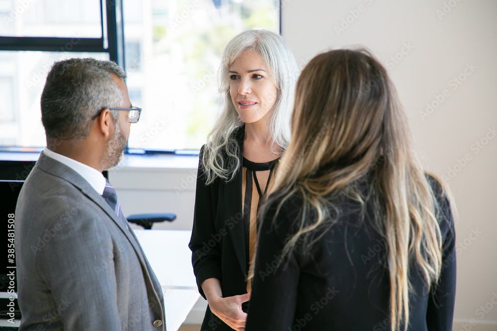Fototapeta premium Company managers wearing suits, standing in office, talking, discussing project. Medium shot. Business communication or briefing concept