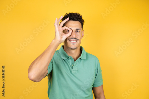 Young handsome man wearing green casual t-shirt over isolated yellow background doing okay gesture shocked with surprised face, eye looking through fingers