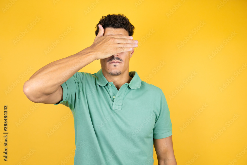 Young handsome man wearing green casual t-shirt over isolated yellow background serious and covering her eyes with her hand Young handsome man wearing green casual t-shirt over isolated yellow background serious and covering her eyes with her hand