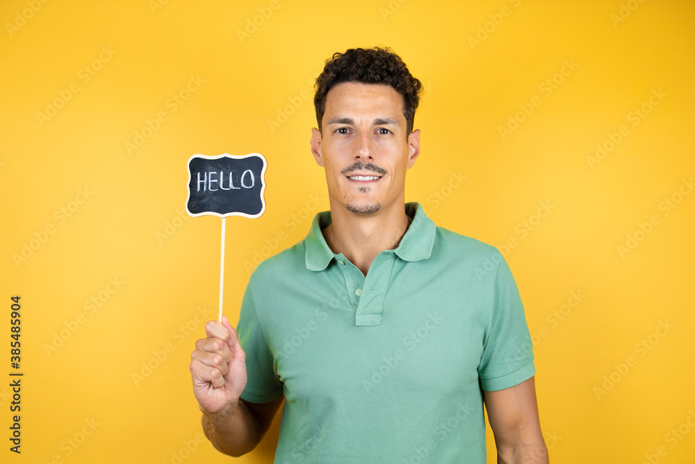 Young handsome man wearing green casual t-shirt over isolated yellow background smiling and holding blackboard with hello word message Young handsome man wearing green casual t-shirt over isolated yellow background smiling and holding blackboard with hello word message