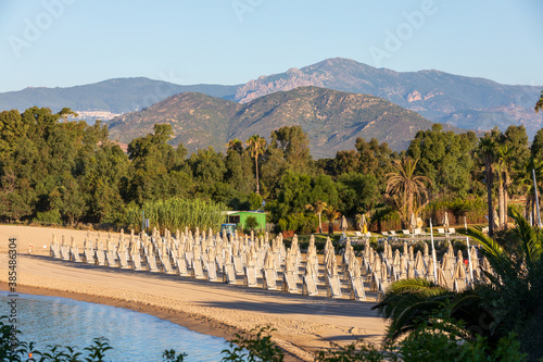 Beach with rows of umbrellas and chairs by the sea in a holiday village during the summer in Italy. Trees and mountains in the background.