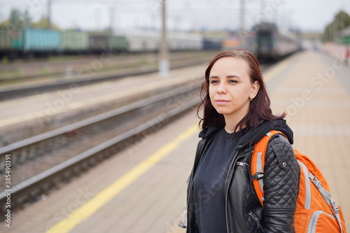 Young woman stands on platform, waiting for train. Portrait of female passenger with backpack on railroad platform in waiting for train ride. Concept of tourism, travel and recreation.