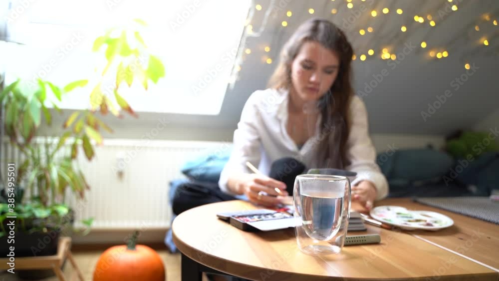 Young woman on sofa near table draws picture in watercolor in notebook Young woman on sofa near table draws picture in watercolor in notebook