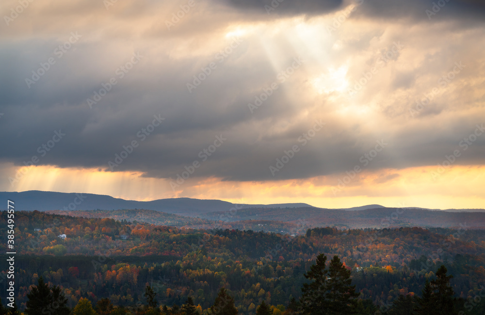 Powerful beams of sunlight breaking through cloudy sky in the afternoon in vermont northeast United States of America during fall shining on a mountain side with vibrant colors of the leaves 
