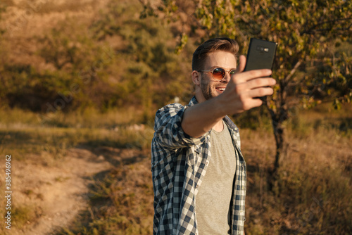 Handsome happy guy taking selfie on cellphone while strolling on nature