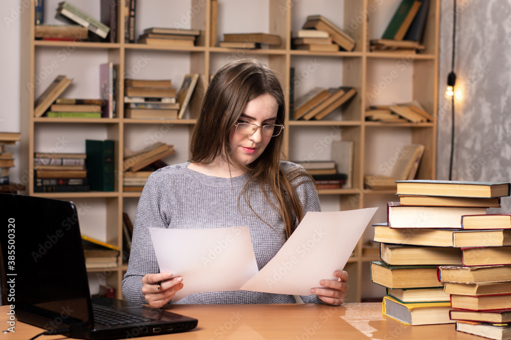 young student prepares for lessons. office worker looks at the documents. a laptop stands on the table and a bookcase in the background. freelancer at work. look at the screen young student prepares for lessons. office worker looks at the documents. a laptop stands on the table and a bookcase in the background. freelancer at work. look at the screen
