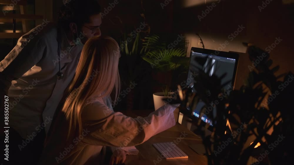 Two cheerful and concentrated doctors using a computer screen researching x ray scan of their patient in dark office room Two cheerful and concentrated doctors using a computer screen researching x ray scan of their patient in dark office room