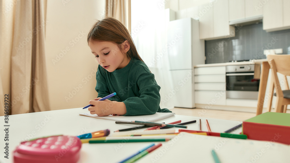 A cute little girl writing with a pencil while sitting at a table having multicolored pencils around the table A cute little girl writing with a pencil while sitting at a table having multicolored pencils around the table