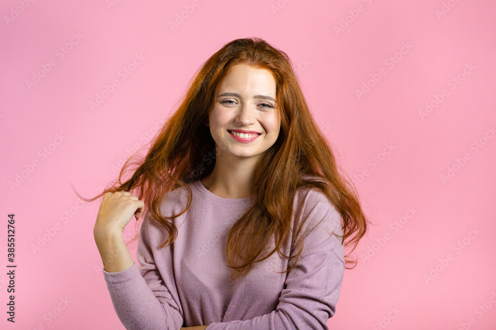 Young woman with long red wavy and shiny hairstyle looking to camera, flirting. Beautiful model girl demonstrate her elegant volume hair. Pink studio background. Young woman with long red wavy and shiny hairstyle looking to camera, flirting. Beautiful model girl demonstrate her elegant volume hair. Pink studio background.