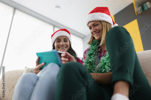 Two happy woman make up wishlist at home. Girlfriends sit on couch and discuss list in notebook.