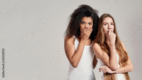 Portrait of two young diverse women in white shirts looking shocked, pensive at camera, covering mouth with hand while posing together isolated over grey background