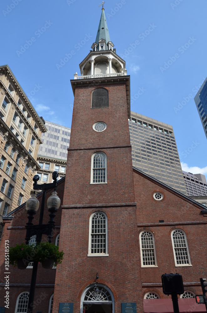 The Old South Meeting House in the middle of Boston with streetlamp in the foreground and some skyscrapers in the background with blue sky