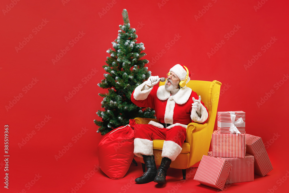 Funny Santa Claus man in Christmas suit sit in armchair with fir tree gifts listen music with headphones sing song on microphone isolated on red background. Happy New Year celebration holiday concept.