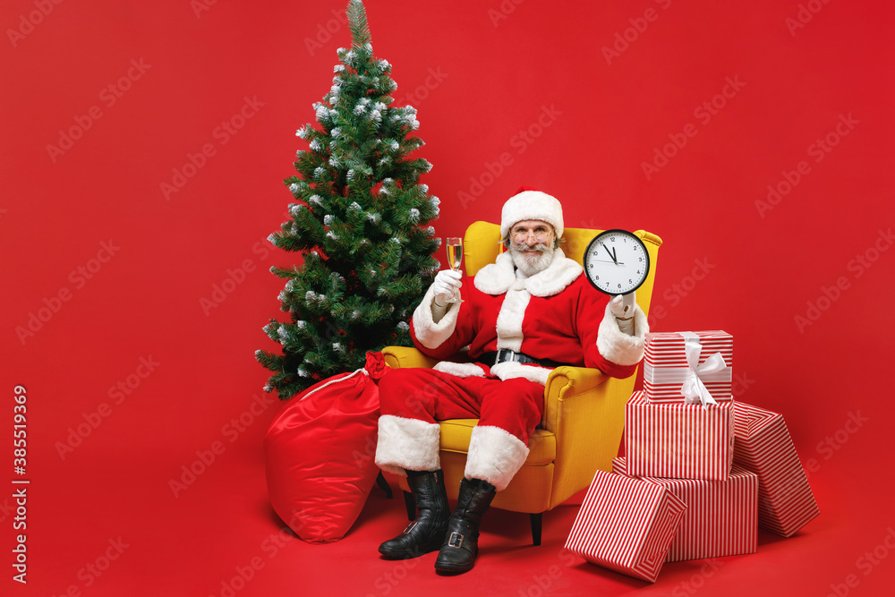 Smiling Santa Claus man in Christmas suit sit in armchair with fir tree presents gifts boxes hold glass of champagne clock isolated on red background. Happy New Year celebration merry holiday concept. Smiling Santa Claus man in Christmas suit sit in armchair with fir tree presents gifts boxes hold glass of champagne clock isolated on red background. Happy New Year celebration merry holiday concept.