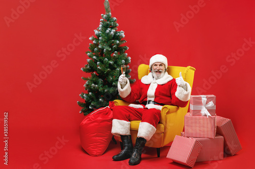 Funny Santa Claus man in Christmas hat suit sit in armchair with fir tree gifts hold glass of champagne showing thumb up isolated on red background. Happy New Year celebration merry holiday concept.