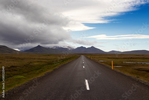 Dramatic Icelandic landscape. Dark asphalt road perspective with wild empty field and mountain range background.