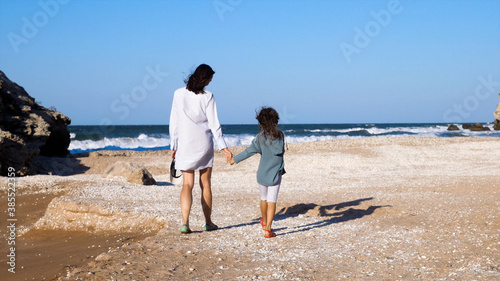 Young attractive woman with little daughter is walking on the beach together, rear view.