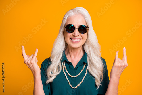 Hard rock Photo of pretty crazy cheerful white haired grandma aged lady showing finger horns metal music lover wear green shirt sun specs necklace isolated bright yellow color background