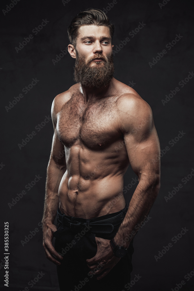 Brutal bearded guy with naked torso and muscular build with hands in pockets posing in dark studio background.