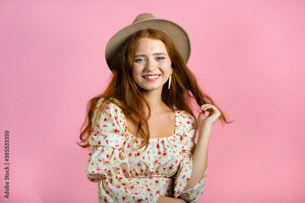 Young woman with long red wavy and shiny hairstyle and hat looking to camera, flirting. Beautiful model girl demonstrate her elegant volume hair. Pink studio background. Young woman with long red wavy and shiny hairstyle and hat looking to camera, flirting. Beautiful model girl demonstrate her elegant volume hair. Pink studio background.