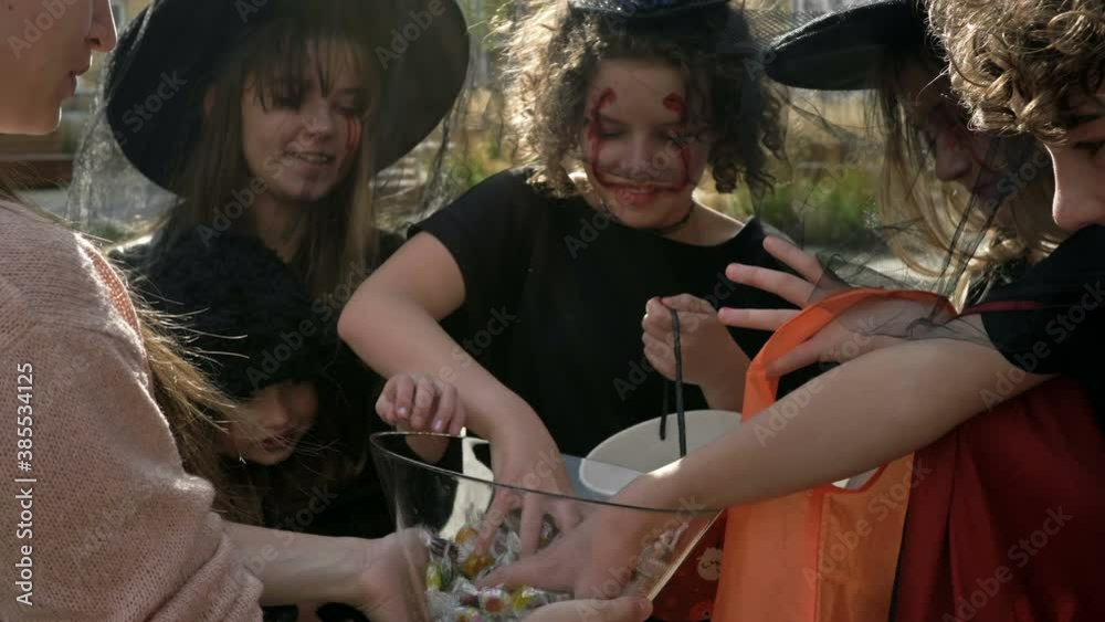 Woman treats sweets to a group of children in black fancy dress with scary makeup on their faces. Halloween traditions.