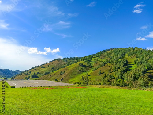 Helipad among mountains with trees, green meadows, blue sky with clouds. Beautiful landscape. Shooting on a sunny warm day