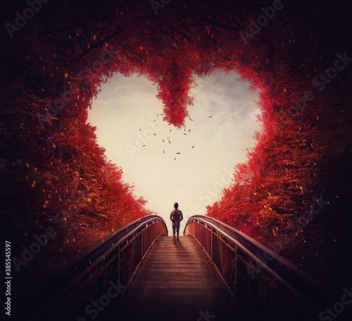 Follow your heart concept. A lone person lost in the autumn forest, found the way out of the woods, as walks a path through the heart shaped trees. Surreal and magic scene, red fall colors. photo