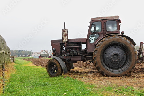 Old wheeled brown diesel tractor on the field with green grass road near wooden fence close up, soil cultivation on an autumn day, countryside agriculture farming landscape