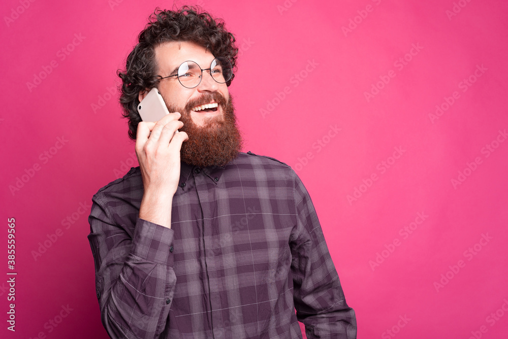 A cheerful bearded young man is talking to his phone and laughing with his glasses on near a pink wall A cheerful bearded young man is talking to his phone and laughing with his glasses on near a pink wall