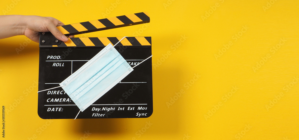 Hand is holding black clapper board or movie slate with face mask. it use in video production and cinema industry on yellow background.